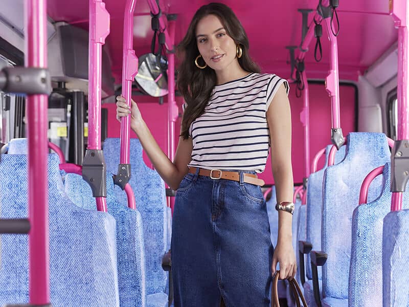 Mulher em ônibus pink com blusa listrada e saia jeans.