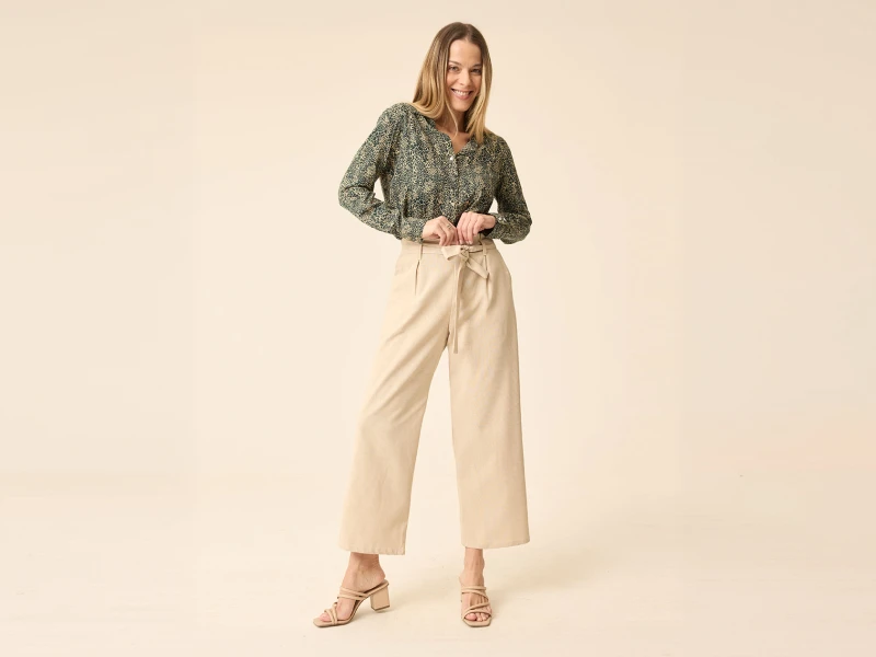 Mulher com camisa padrão verde, calças beges de perna larga e sandálias de salto alto.