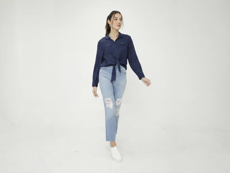 Mulher com camisa azul-marinho atada, calças de ganga claras com rotos e sapatilhas brancas.