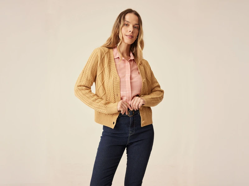 Mulher com casaco de malha amarelo torrado, camisa rosa e calças de ganga azuis escuras.