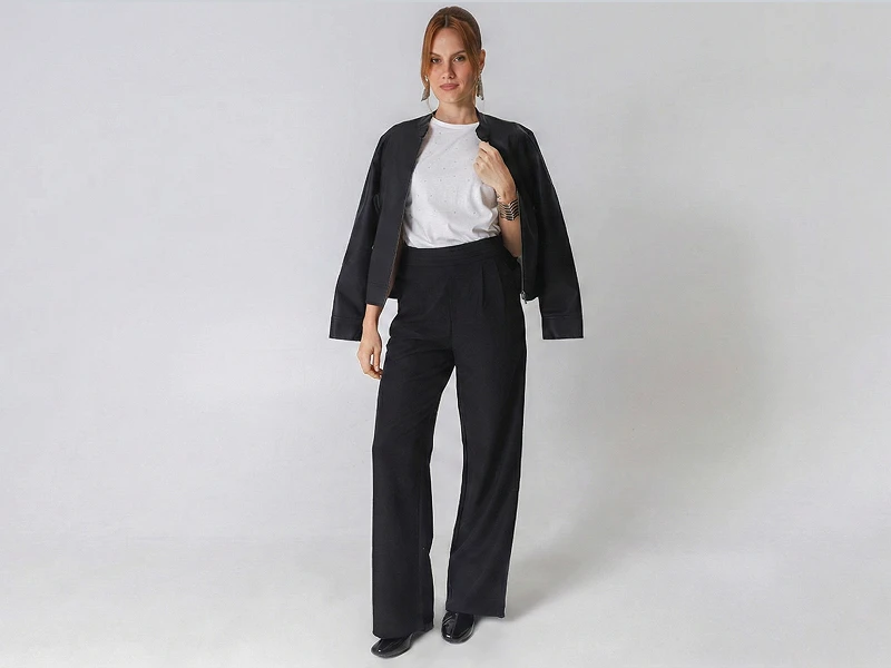 Mulher com casaco preto sobre os ombros, t-shirt branca e calças pretas largas em fundo branco.