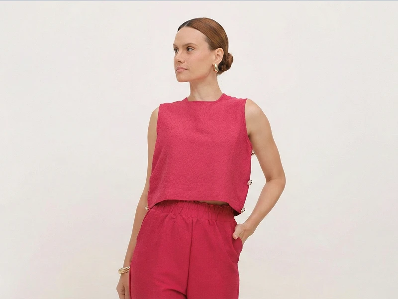 Mulher com conjunto fúcsia de top sem mangas e calças de cintura subida sobre fundo neutro.