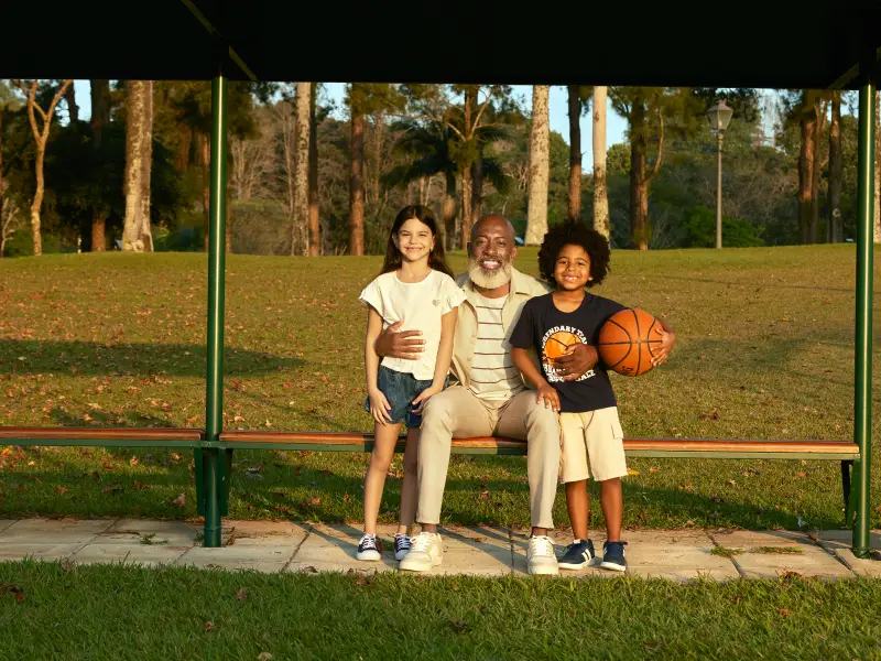 Avô e netos sorriem num banco de parque ao pôr do sol, com o rapaz a segurar uma bola de basquetebol.