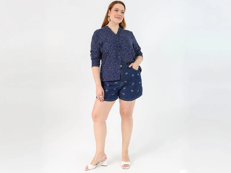 Mulher ruiva veste camisa azul marinho às bolinhas e calções de ganga com laços bordados.