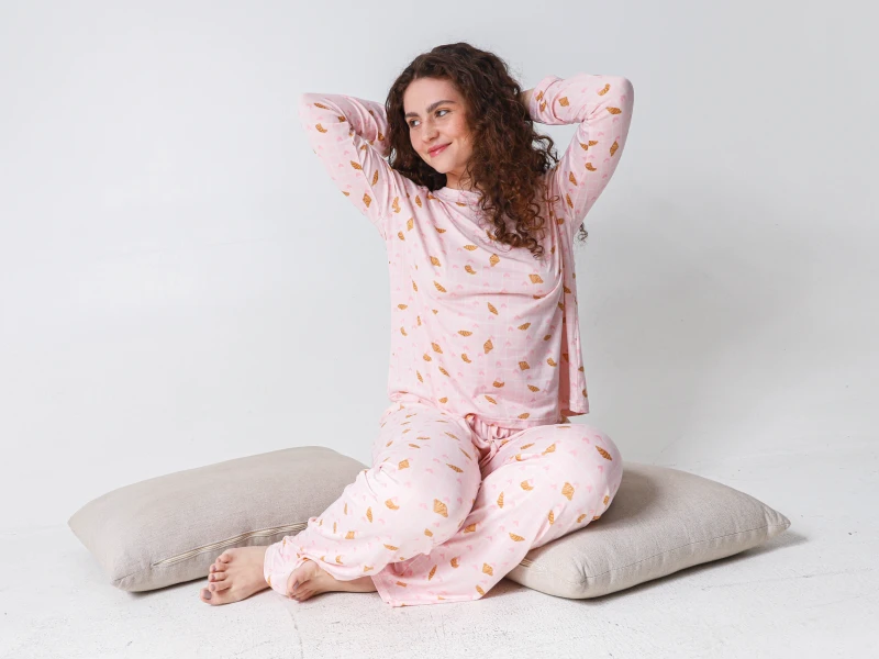 Mulher com pijama cor-de-rosa de mangas compridas e padrão de croissants, sentada no chão junto a almofadas.