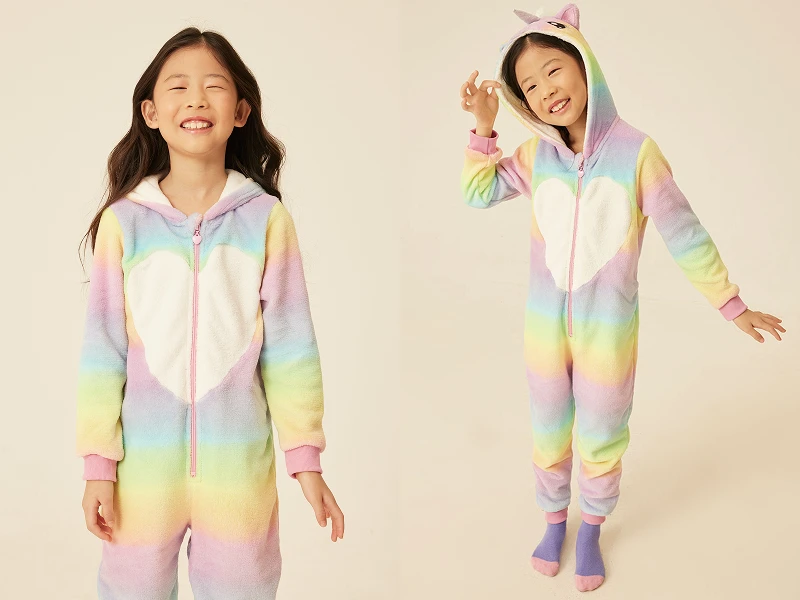 Menina sorridente com pijama macacão arco-íris, capuz de unicórnio e detalhe de coração branco.