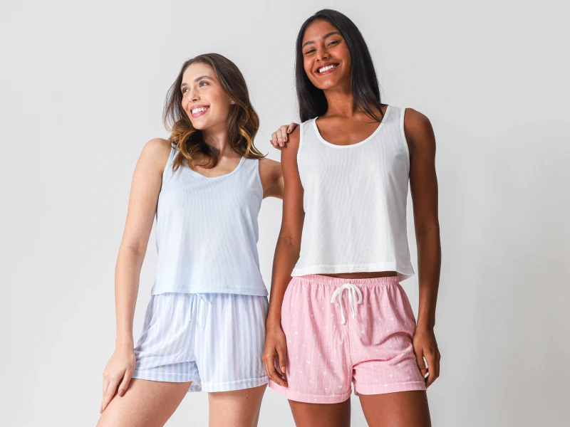 Duas mulheres sorridentes com pijamas de verão: conjunto azul às riscas e conjunto rosa com padrão.