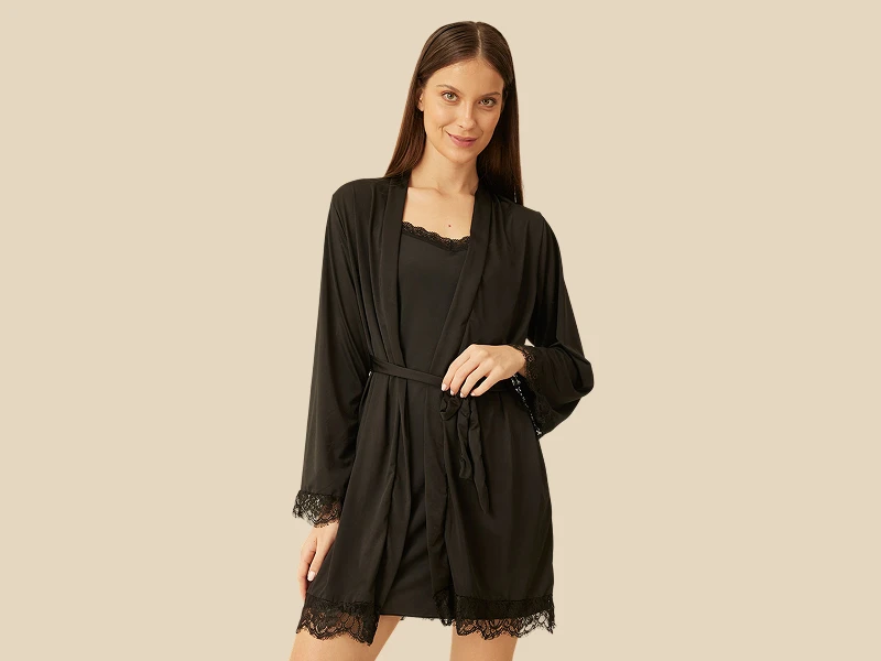 Mulher a posar com robe preto de cetim e renda sobre camisola de noite preta em fundo bege.