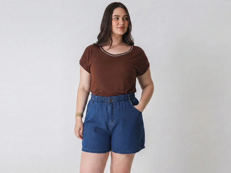 Mulher veste t-shirt castanha e calções de ganga azuis de cintura elástica sobre fundo branco.