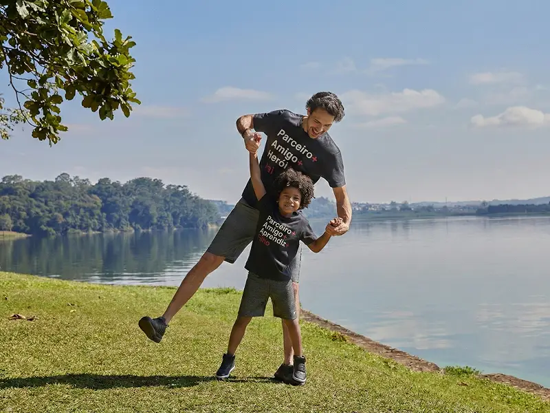 Pai e filho com t-shirts personalizadas brincam sorridentes junto a um lago num dia ensolarado.