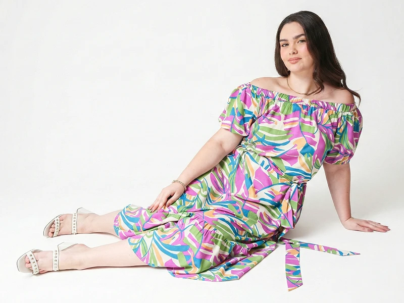 Mulher plus-size com vestido colorido de padrão tropical e ombros à mostra, sentada num fundo branco.