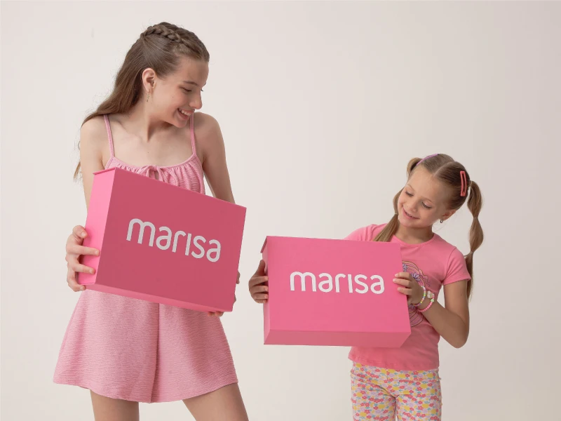 Duas meninas sorridentes com roupa cor-de-rosa a segurar caixas de presente da marca Marisa.