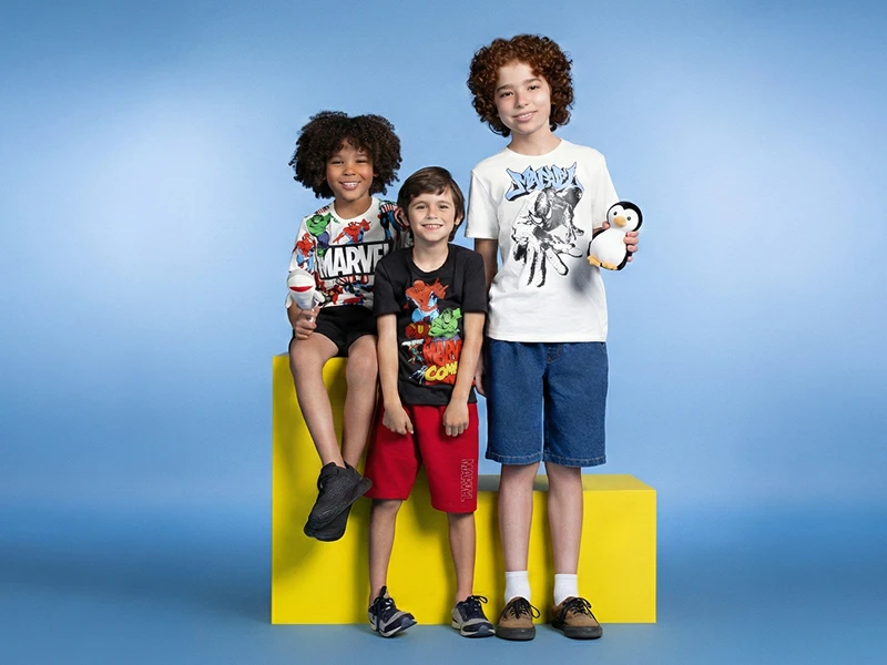 Três meninos com t-shirts da Marvel e calções casuais posam em blocos amarelos sobre fundo azul.