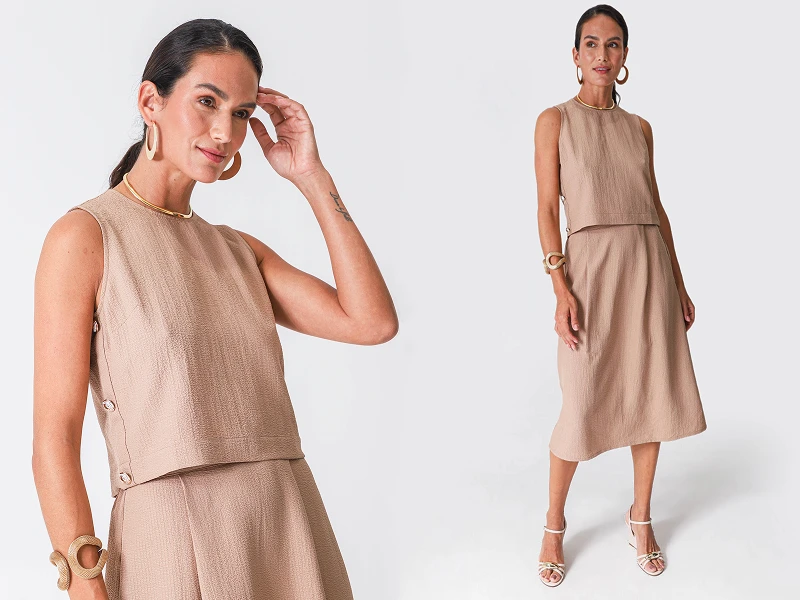 Mulher com conjunto bege de top sem mangas e saia midi com acessórios dourados em fundo branco.