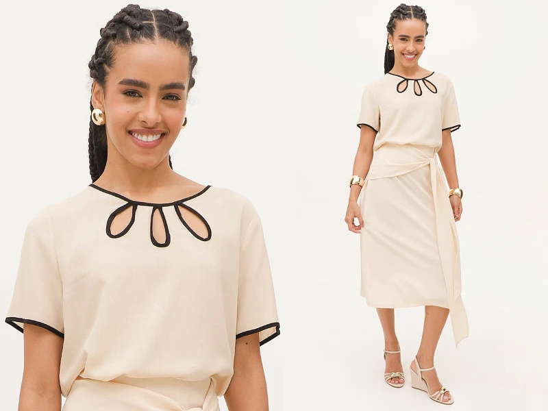 Mulher sorridente com tranças usa vestido bege midi com pormenores pretos no decote e cintura com laço.
