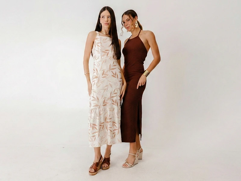 Duas mulheres com vestidos compridos: um branco com padrão de folhas e outro castanho com decote halter e racha.